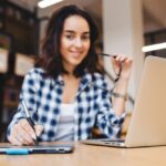 Imagem elegante moderna de jovem morena inteligente trabalhando com o laptop na mesa na biblioteca sorrindo brincando de óculos escuros grande sucesso estudante trabalhador